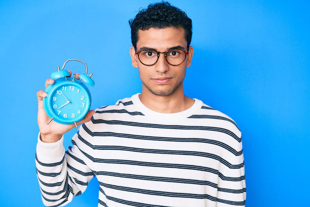 Young handsome hispanic man holding alarm clock thinking attitude and sober expression looking self confident  - Photo, Image