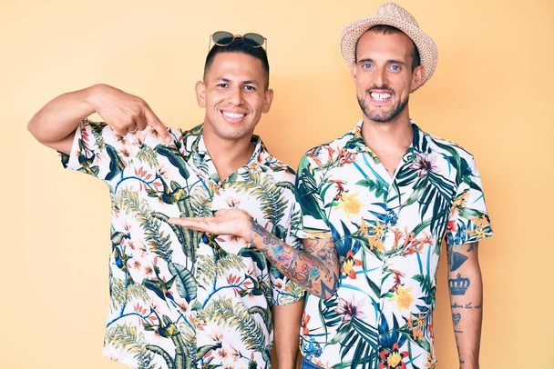 Jeune couple gay de deux hommes portant chapeau d'été et chemise hawaïenne étonné et souriant à la caméra tout en présentant avec la main et pointant du doigt.  - Photo, image