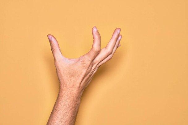 Hand van blanke jongeman met vingers over geïsoleerde gele achtergrond plukken en nemen onzichtbare ding, houden object met vingers tonen ruimte - Foto, afbeelding