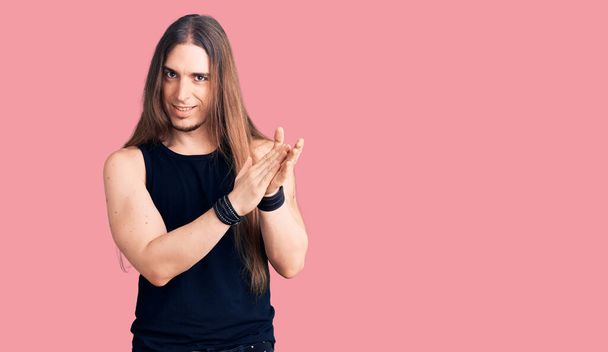 Young adult man with long hair wearing goth style with black clothes clapping and applauding happy and joyful, smiling proud hands together  - Foto, Imagem