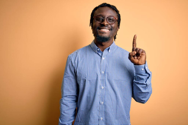 Junger gutaussehender afrikanisch-amerikanischer Mann, der Hemd und Brille über gelbem Hintergrund trägt und mit Finger Nummer eins zeigt, während er selbstbewusst und glücklich lächelt. - Foto, Bild