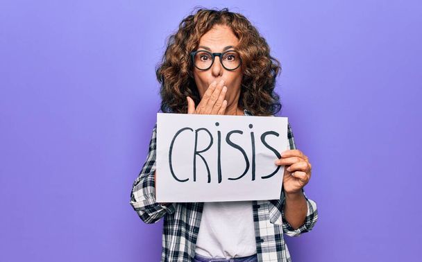 Middle age beautiful woman holding paper with crisis message over purple background covering mouth with hand, shocked and afraid for mistake. Surprised expression - Foto, Bild