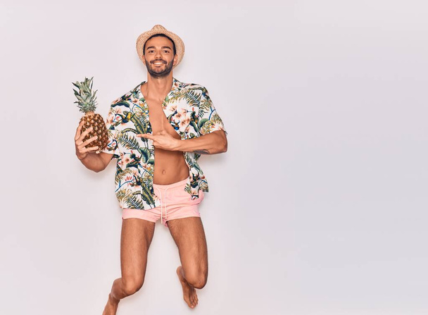 Young handsome hispanic man on vacation wearing swimwear, floral shirt and hat smiling happy. Jumping with smile on face holding pineapple over isolated white background - Photo, Image