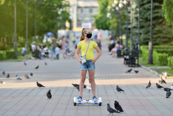 Girl child in a black protective mask rides on a hoverboard past pigeons in a park during the COVID-19 pandemic - Fotoğraf, Görsel