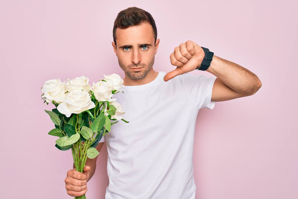 Fiatal jóképű férfi, kék szemekkel, rózsaszín háttér felett virágcsokrot tartva dühös arccal, negatív jel, ami nem tetszik hüvelykujjal lefelé, elutasítás koncepció - Fotó, kép