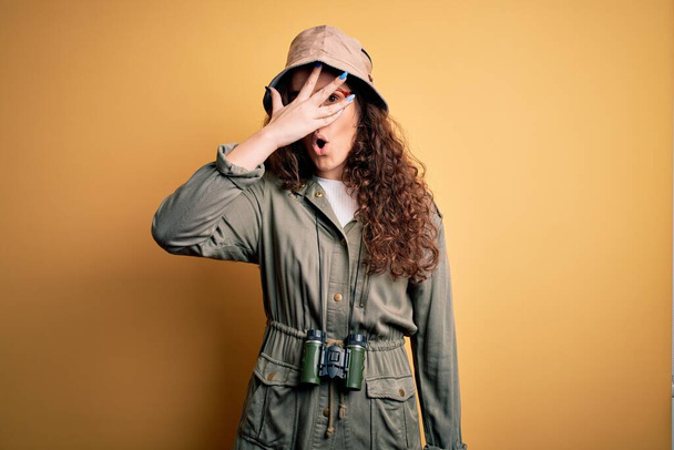 Young beautiful tourist woman on vacation wearing explorer hat and binoculars peeking in shock covering face and eyes with hand, looking through fingers with embarrassed expression. - Photo, Image