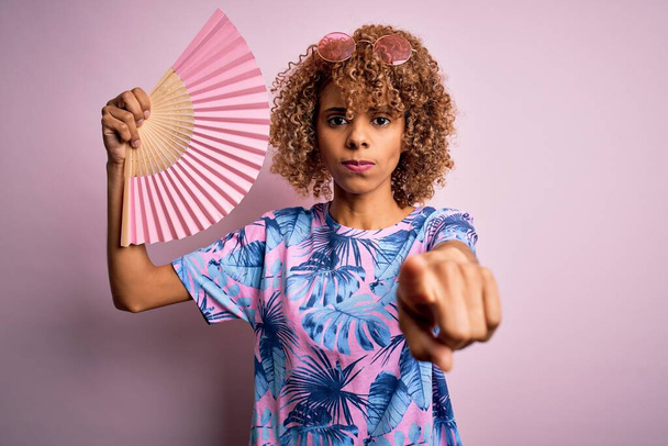 Joven mujer rizada afroamericana de vacaciones usando ventilador de mano sobre fondo rosa aislado apuntando con el dedo a la cámara y a usted, signo de mano, gesto positivo y seguro desde el frente
 - Foto, Imagen