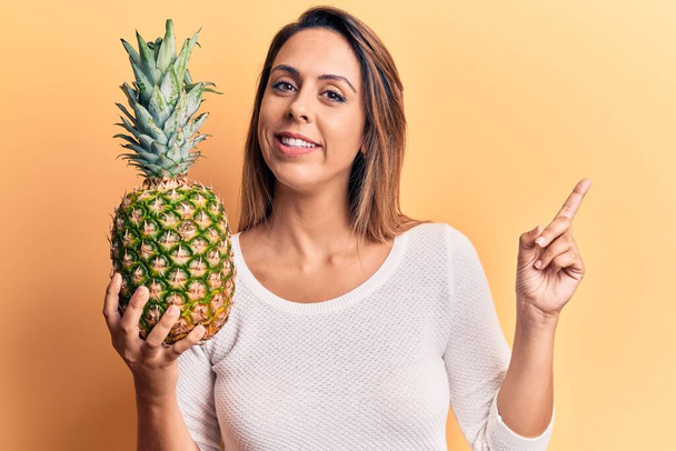 Young beautiful woman holding pineapple smiling happy pointing with hand and finger to the side  - Photo, Image