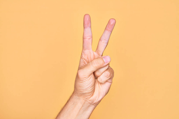 ręka kaukaski młody człowiek pokazując palce na odizolowanym żółtym tle licząc numer 2 pokazując dwa palce, gestykulując zwycięstwo i symbol zwycięzca - Zdjęcie, obraz