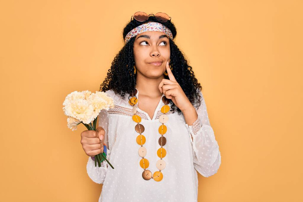 Młoda, afrykańska hipisowska, kręcona kobieta w okularach przeciwsłonecznych, trzymająca bukiet kwiatów, poważnie myśląca o pytaniu, bardzo zdezorientowany pomysł. - Zdjęcie, obraz