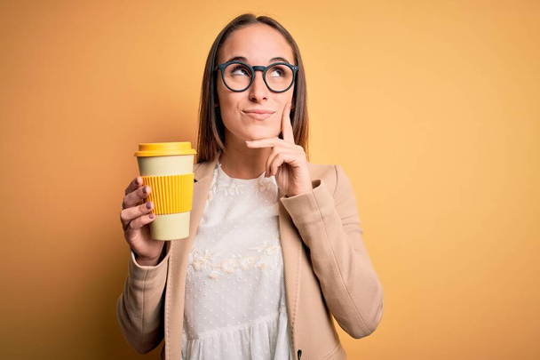 Junge schöne Geschäftsfrau trinkt Tasse Kaffee zum Mitnehmen über gelbem Hintergrund ernstes Gesicht über Frage nachdenkend, sehr verwirrte Idee - Foto, Bild