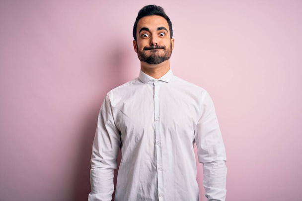 Young handsome man with beard wearing casual shirt standing over pink background puffing cheeks with funny face. Mouth inflated with air, crazy expression. - Photo, Image