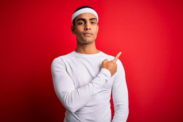 Young handsome african american sportsman wearing sportswear over red background Pointing with hand finger to the side showing advertisement, serious and calm face - 写真・画像