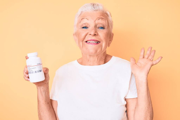 Senior beautiful woman with blue eyes and grey hair holding pills celebrating victory with happy smile and winner expression with raised hands  - 写真・画像