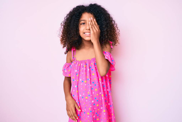 Niño afroamericano con el pelo rizado usando vestido casual cubriendo un ojo con la mano, sonrisa confiada en la cara y emoción sorpresa
.  - Foto, Imagen