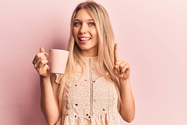 Young beautiful blonde woman holding coffee smiling with an idea or question pointing finger with happy face, number one  - Foto, Imagem
