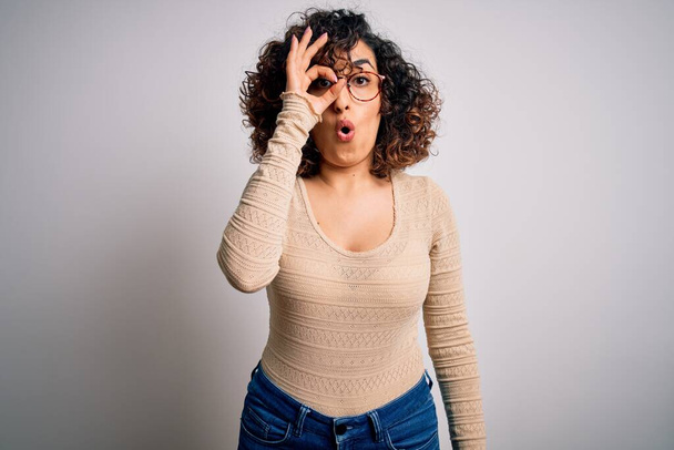 Young beautiful curly arab woman wearing casual t-shirt and glasses over white background doing ok gesture shocked with surprised face, eye looking through fingers. Unbelieving expression. - Fotografie, Obrázek