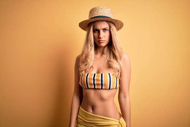 Junge schöne blonde Frau im Urlaub mit Bikini und Hut über gelbem Hintergrund skeptisch und nervös, Stirnrunzeln aufgeregt wegen des Problems. Negative Person. - Foto, Bild