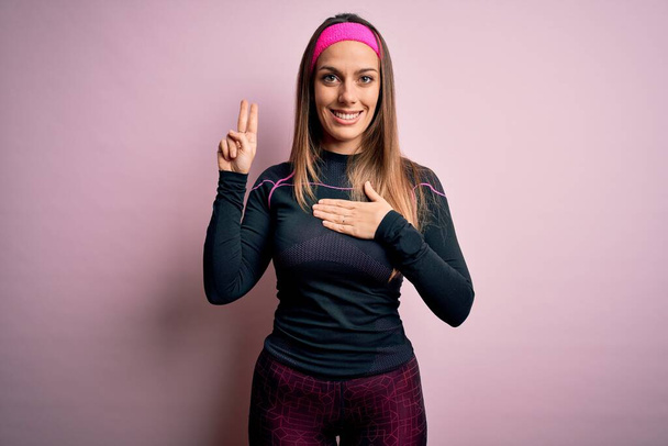 Fiatal szőke fitness nő visel sport edzőruha felett elszigetelt háttér mosolygós káromkodás kezét a mellkas és az ujjak fel, hogy hűségesküt - Fotó, kép