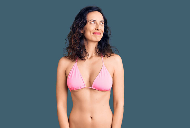 Junge schöne hispanische Frau im Bikini lächelt zur Seite und starrt nachdenklich weg.  - Foto, Bild