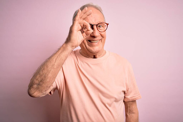 Hombre mayor de pelo gris con gafas de pie sobre fondo aislado rosa haciendo buen gesto con la mano sonriendo, los ojos mirando a través de los dedos con la cara feliz
. - Foto, imagen