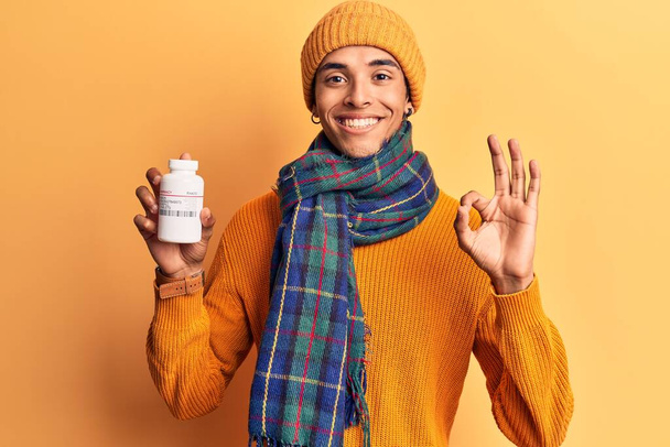 Young african amercian man wearing winter clothes holding pills doing ok sign with fingers, smiling friendly gesturing excellent symbol  - Foto, Bild