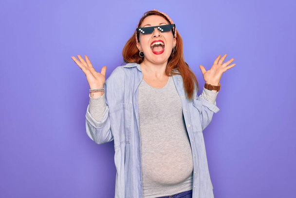 Young redhead pregnant woman expecting baby wearing funny thug life sunglasses celebrating mad and crazy for success with arms raised and closed eyes screaming excited. Winner concept - Foto, Bild