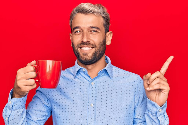 Fiatal, jóképű, szakállas szőke férfi, vörös bögre kávét iszik elszigetelt háttér felett, mosolyogva, boldogan mutogatva, kezével és ujjával oldalra. - Fotó, kép