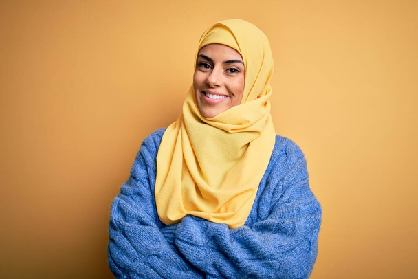 Fiatal gyönyörű barna muszlim nő visel arab hidzsáb felett elszigetelt sárga háttér boldog arc mosolyogva keresztbe karok nézi a kamerát. Pozitív személy. - Fotó, kép