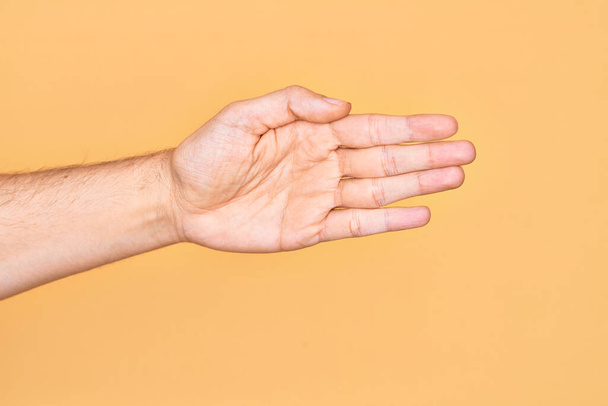 Main de jeune homme caucasien montrant les doigts sur fond jaune isolé étirant et atteignant avec la main ouverte pour la poignée de main, montrant la paume - Photo, image