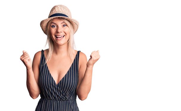 Young beautiful blonde woman wearing summer hat and dress celebrating surprised and amazed for success with arms raised and open eyes. winner concept.  - Photo, image