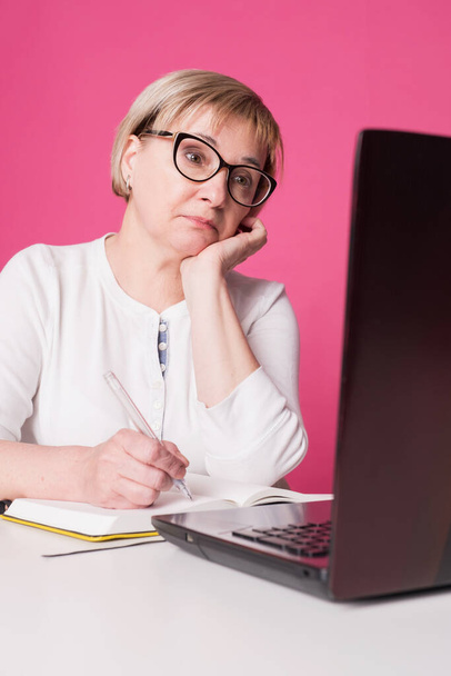 Old woman in her 60s works on computer, wearing headphohes. Laptop on white table and Pink background. She sits thoughtful, unhappy, a bit sad - Foto, Imagen