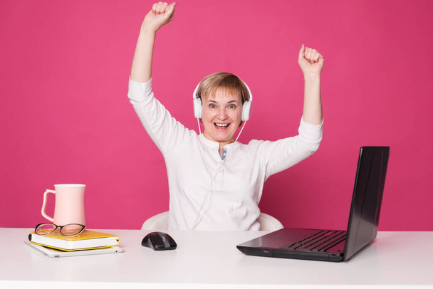 Old woman in her 60s works on computer, wearing headphohes. Laptop on white table and Pink background. She make happy winning gestures - Photo, Image