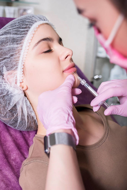 Cosmetoloog arts doet een injectieprocedure bij een vrouw in haar kliniek. Ze draagt gloed en masker en houdt de spuit vast met een naald. - Foto, afbeelding