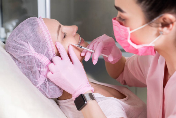 Cosmetologist doctor do injection procedure to a woman in her clinic. She wear pink glowes and mask and holds syringe with a needle. - Photo, Image