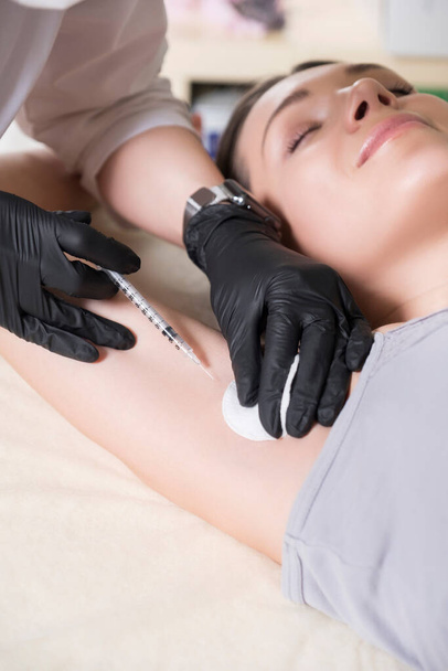Kosmetologe Arzt machen Injektionsverfahren zu einer Frau in ihrer Klinik. Sie trägt schwarze Handschuhe und Maske und hält Spritze mit Nadel. - Foto, Bild