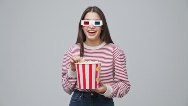 Smiling attractive brunette woman in 3D glasses eating popcorn and pointing at the camera over grey background - Záběry, video