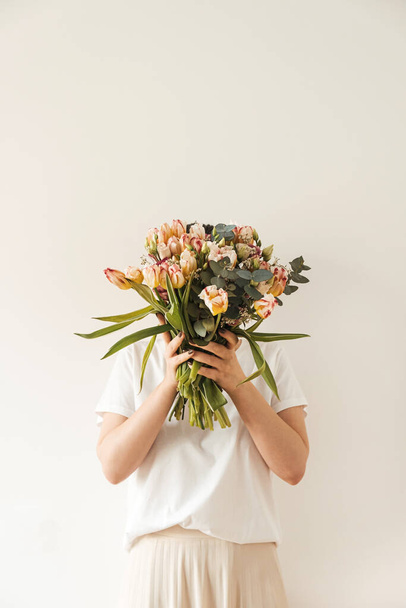 Mujer bonita joven en blusa blanca con flores de tulipán ramo en las manos contra la pared blanca. Celebración festiva concepto floral - Foto, Imagen