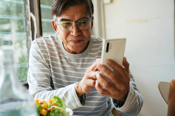 Старий чоловік в окулярах посміхається, приймаючи відеодзвінок зі своєю дитиною по смартфону
. - Фото, зображення