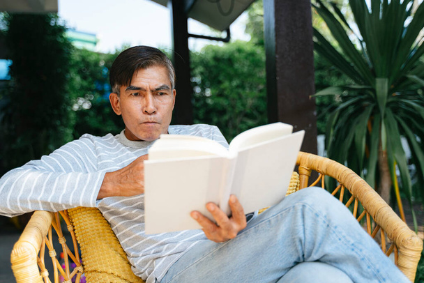 Старик читает книгу и сринк воду, сидя на плетеной скамейке во дворе
. - Фото, изображение