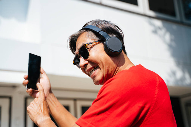 Alter Mann in rotem T-Shirt genießt die Musik aus mobiler Anwendung bei sonnigem Tag zu Hause. - Foto, Bild