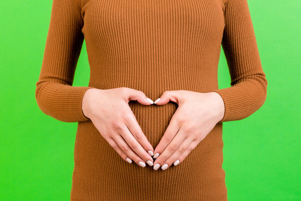 Primer plano del vientre de la mujer embarazada con el corazón hecho de dedos en el fondo verde. Mamá lleva un vestido marrón. Concepto de maternidad. Amor al futuro bebé. Copiar espacio
. - Foto, Imagen
