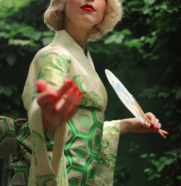 Blondýnka s červenými rty a v zeleném kimonu drží bílý vějíř s modrými kresbami v rukou a gesto volá na sebe - Fotografie, Obrázek