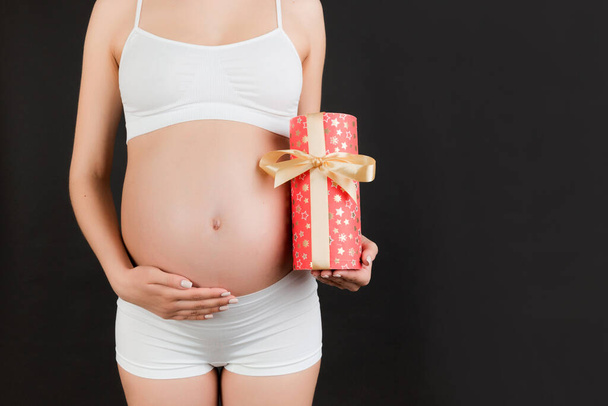 Beyaz iç çamaşırlı hamile bir kadının hediye kutusunu tutarken ve mavi arka planda karnına dokunurken görüntüsü kesilmiş. Bir kız bebek bekliyorum. Boşluğu kopyala. - Fotoğraf, Görsel