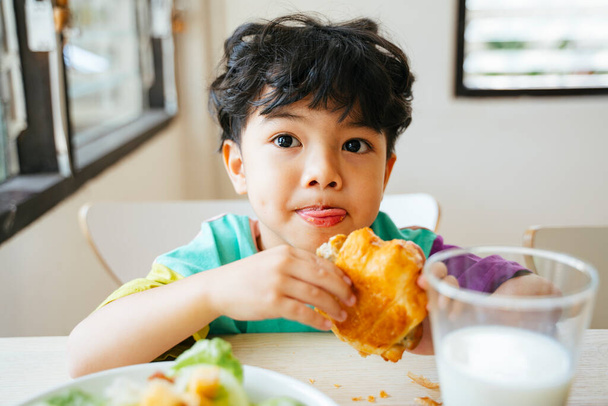 Kleiner Junge hält das Wurst-Croissant in den Händen mit einem glücklichen Lächeln im Gesicht. - Foto, Bild