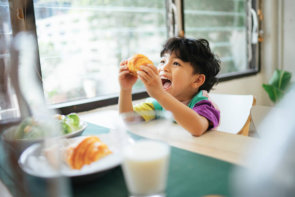 Niño pequeño sostiene el croissant salchicha en sus manos con sonrisa de felicidad en su cara
. - Foto, imagen