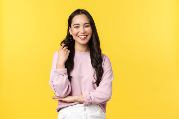 People emotions, lifestyle and fashion concept. Laughing asian girl, casualy talking, touching hair strand and smiling at camera, pleased with new haircut, enjoying summer, stand yellow background - Photo, image