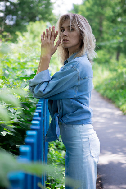 A girl with blond hair and a blue denim jacket is leaning against a blue fence. - Fotografie, Obrázek