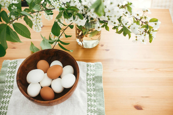 Happy Easter zero waste concept. Natural Easter eggs white and brown in stylish wooden bowl on linen fabric on background of cherry flowers on green branches. Organic eggs - Photo, image