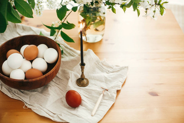Natural Easter eggs in wooden bowl and vintage candle on linen fabric on background of cherry flowers and green leaves on rural  table. Happy Easter. Organic eggs, zero waste holiday - Photo, image
